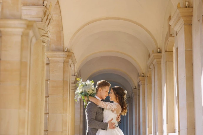 Couple de mariés debout l'un face à l'autre sous les arcades en pierre d'un bâtiment historique, baigné de lumière naturelle.
