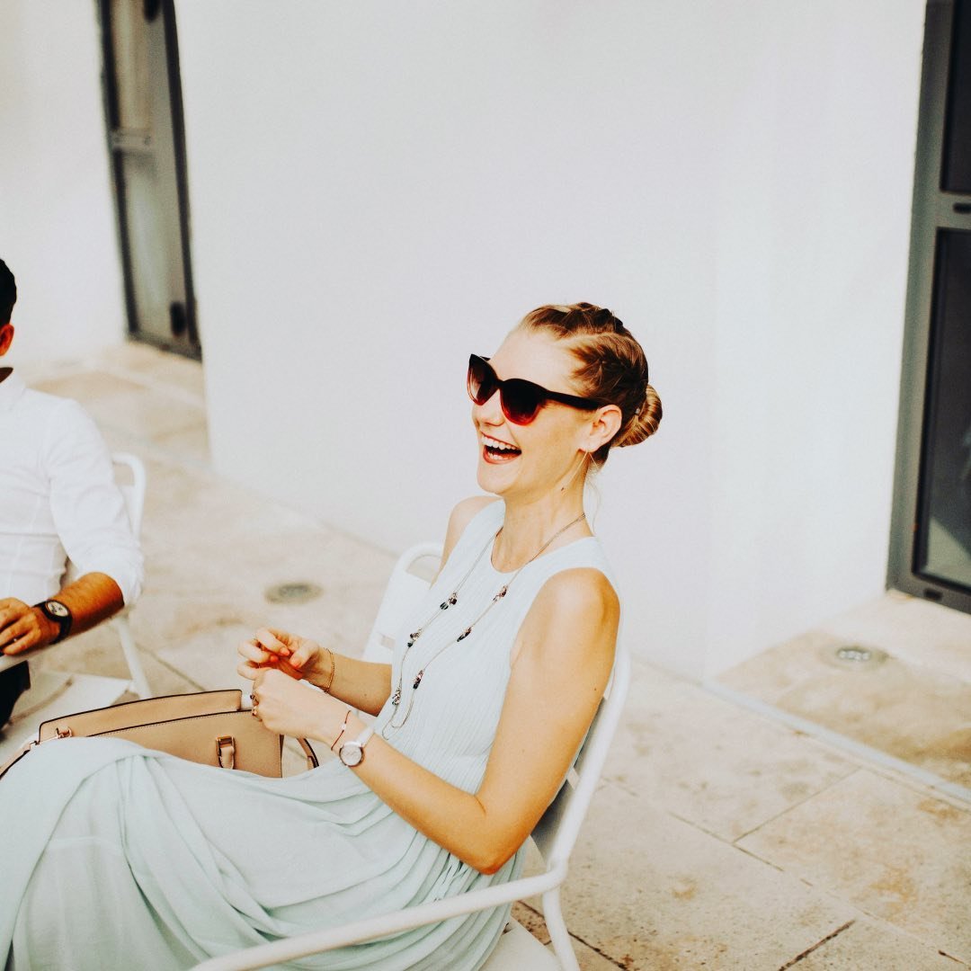 Portrait spontané d'une jeune femme riant aux éclats, portant des lunettes de soleil rouges lors d'un cocktail de mariage en extérieur.